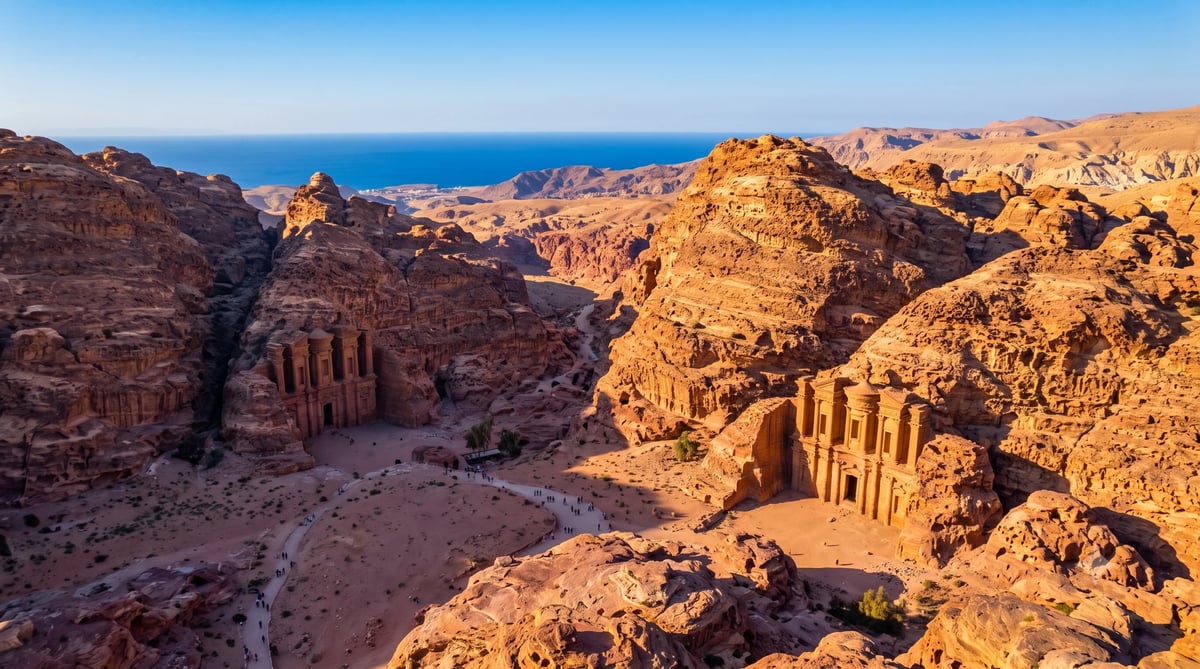 Ancient carved stone structures nestled in a rocky desert valley with the blue sea visible in the distance