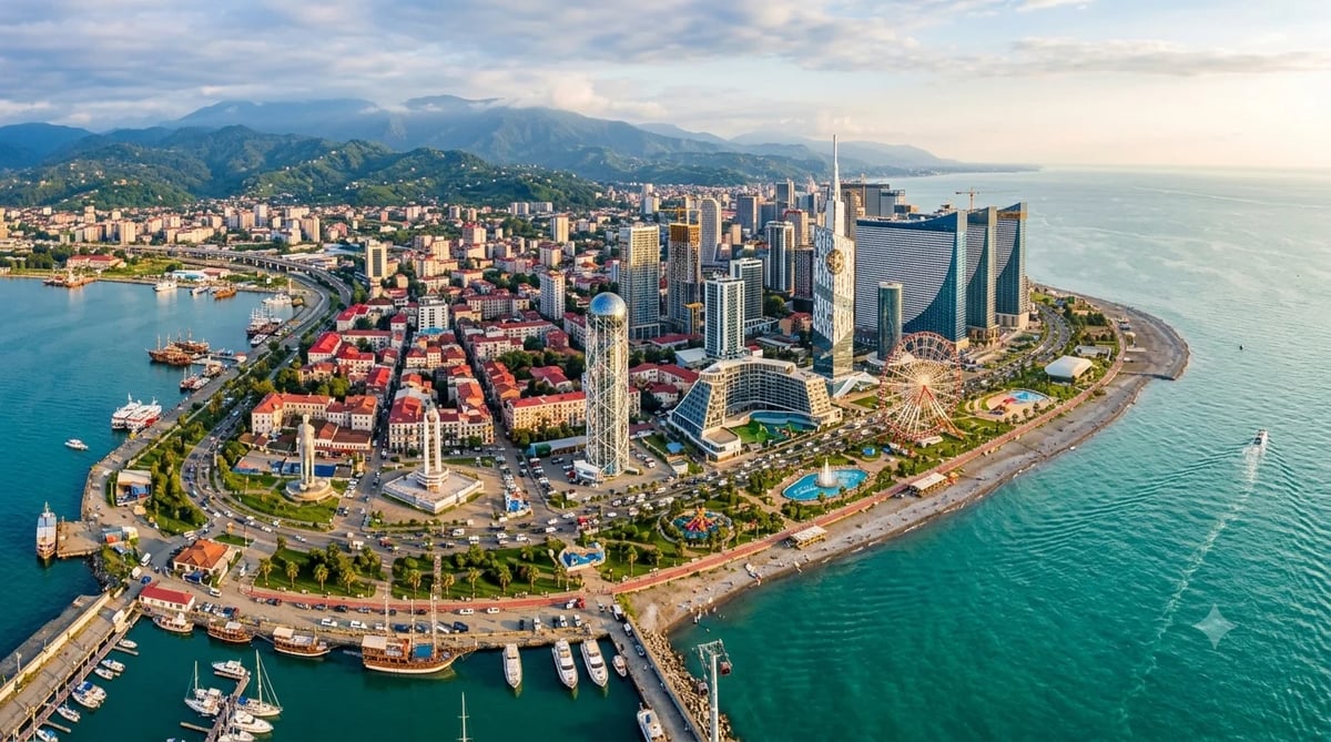 Aerial view of Batumi's coastal skyline with modern skyscrapers, harbor, Ferris wheel, and the Caucasus Mountains in the background